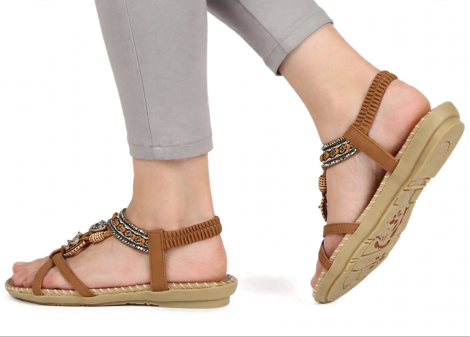 A pair of women's flat sandals with brown straps, woven trim, and metallic embellishments, shown on a model's feet against a white background.